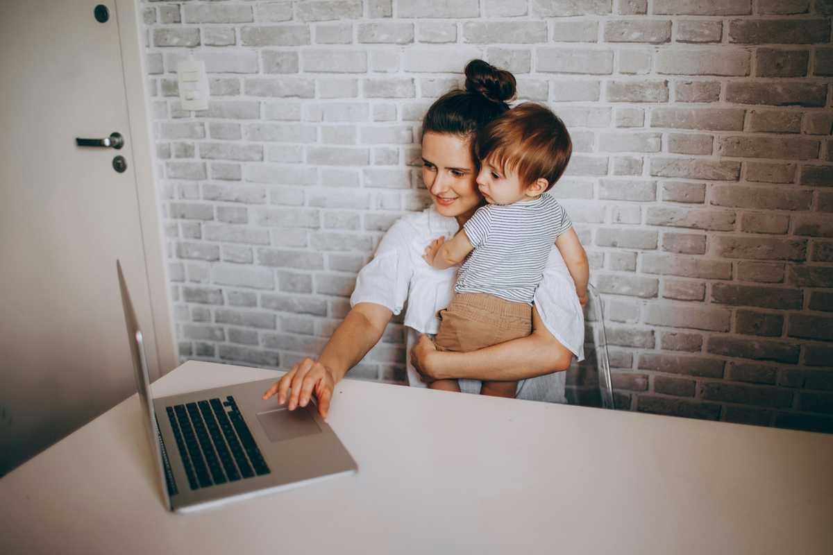 A mother holding her toddler while using a laptop at home